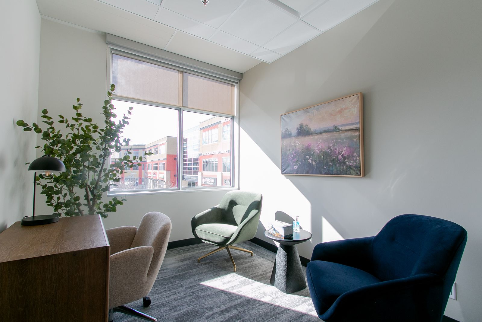 A sunlit studio room at Clinic Studios with a window, a green chair, a navy chair, a tall plant and a framed landscape painting