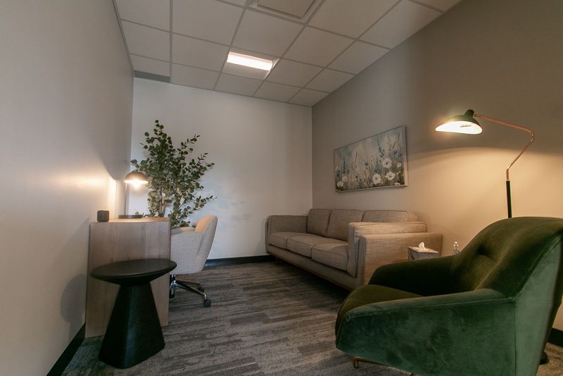 A therapy room at Clinic Studios in lamplight: a sofa, a green velvet armchair, a floor lamp and a desk