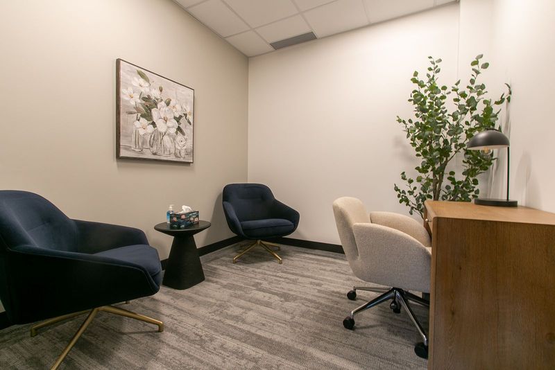 A studio room with two navy armchairs, a desk and chair, a tall plant and framed floral art